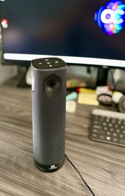 Black cylindrical wireless speaker with fabric cover on a wooden desk, monitor and keyboard in the background.