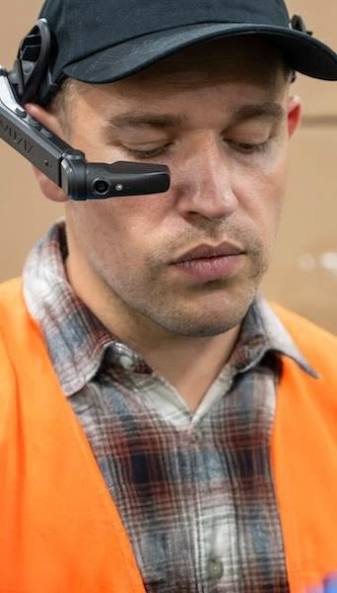 Close-up of a man wearing a black cap and orange safety vest with a headset microphone, looking down.