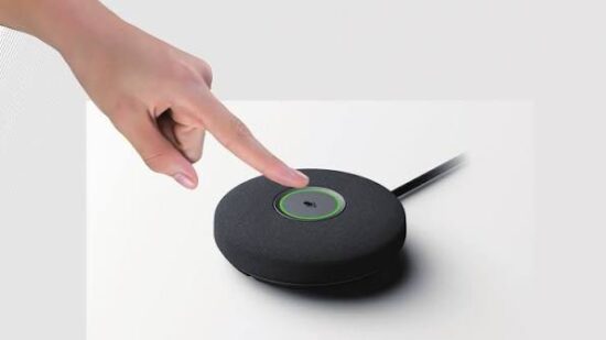 Hand pressing the top button on a black circular smart speaker placed on a white surface.
